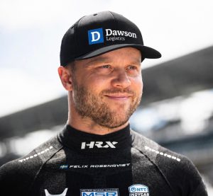 Portrait of a man in a black Dawson Logistics cap and racing suit, smiling outdoors.