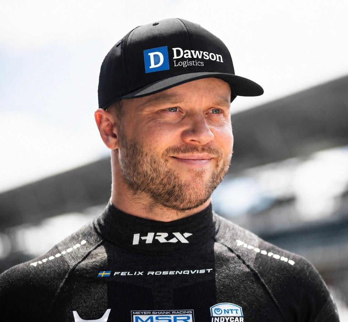 Portrait of a man in a black Dawson Logistics cap and racing suit, smiling outdoors.